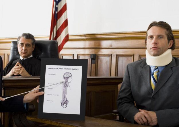 A lawyer pointing at an exhibit of a spinal chord diagram during a trial for an injury case with the injured party on the bench and the judge looking over